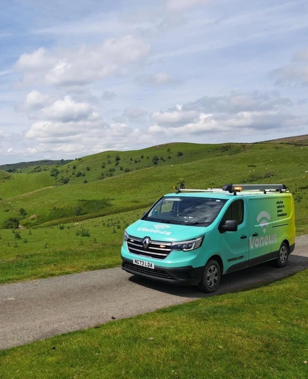 Turquoise Voneus broadband van on a countryside lane beside grassy hills beneath a partly cloudy sky in Gloucestershire.
