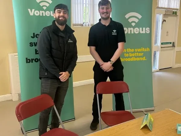 Two Voneus representatives in a community hall with green broadband banners and leaflets displayed on a table in East Devon.