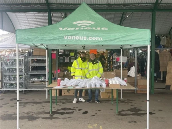 Two Voneus team members in high-visibility jackets at a promotional stand beneath a green branded gazebo inside a market in Somerset.