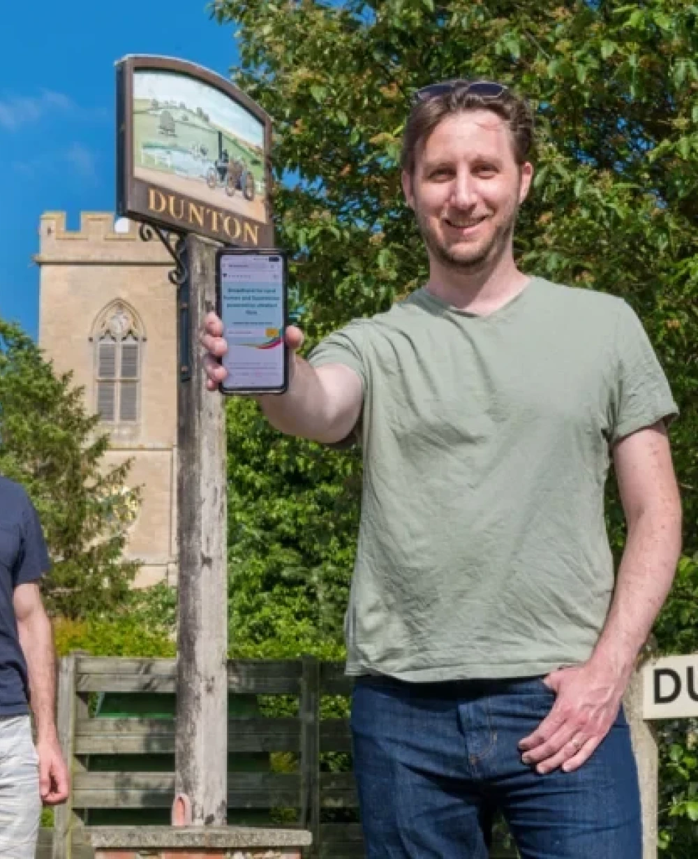 Two local residents using their smartphones beside the Dunton village sign, highlighting rural broadband connectivity near Biggleswade and Potton by Voneus
